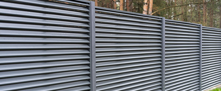 Gray metal louvered privacy fence panels along wooded property edge, modern horizontal slat design