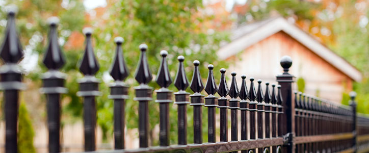 Black wrought-iron fence with decorative finials in front of a suburban house, autumn foliage blurred behind