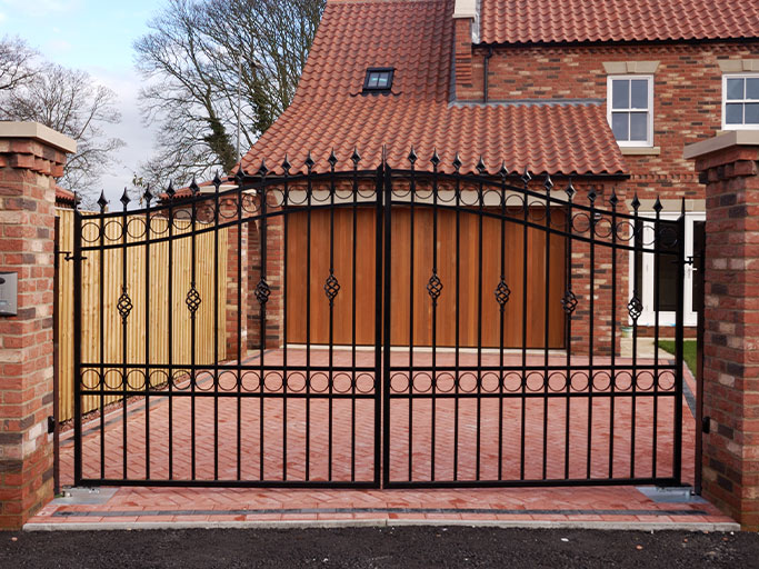 Wrought iron driveway gate in front of brick house with wooden garage doors and red tiled roof