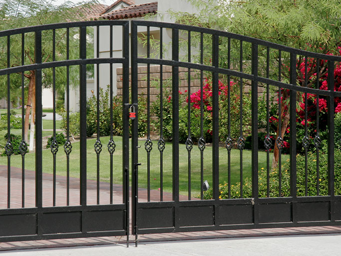 Black wrought-iron driveway gate in front of a suburban house with lawn and red flowering shrubs.