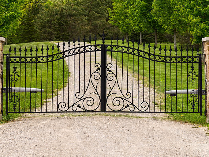 Ornate black wrought-iron driveway gate with decorative scrollwork across gravel road, flanked by green lawn and trees.