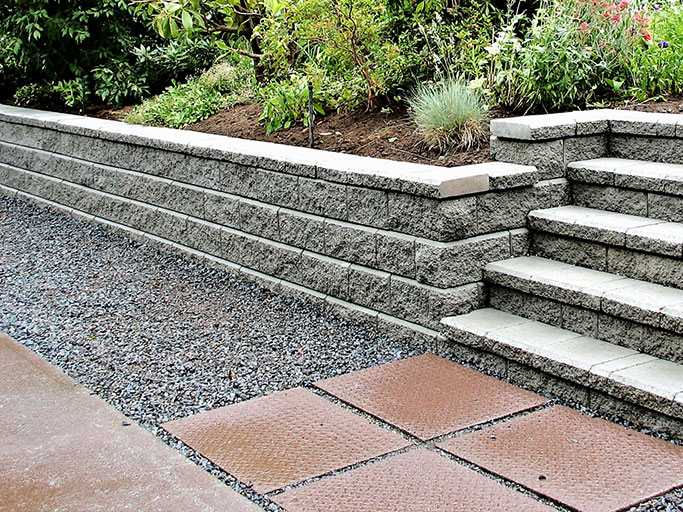 Stone retaining wall with concrete steps, gravel path and square reddish pavers leading to a planted garden bed.