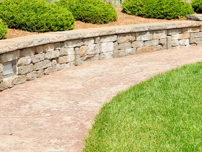 Curved stamped concrete walkway by stone retaining wall, manicured lawn and evergreen shrubs in landscaped yard.