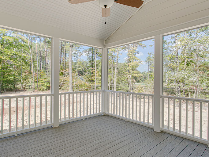 Bright screened porch with ceiling fan, white railings and gray deck flooring overlooking a wooded backyard.