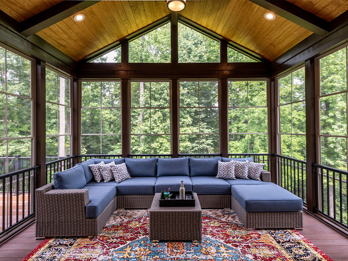 Screened porch with wicker sectional sofa with blue cushions, patterned rug, coffee table, and lush forest views.