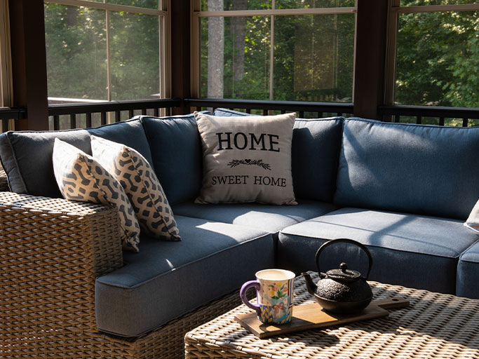 Sunlit enclosed porch with blue sectional sofa, Home Sweet Home pillow, wicker coffee table with teapot and mug.