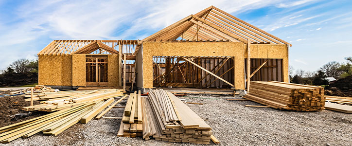 Partially framed suburban house under construction with stacked lumber and gravel drive.
