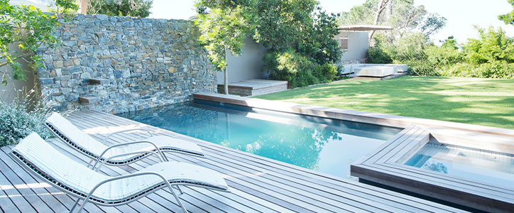 Modern backyard pool with wooden deck, two lounge chairs, stone accent wall, spa, and manicured lawn.