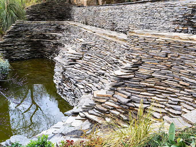 Curving layered dry-stone terraces forming a stepped retaining wall around a reflective garden pond with ornamental grasses