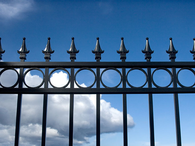 Ornate black wrought-iron fence with spiked finials and circular motifs silhouetted against a blue sky with clouds