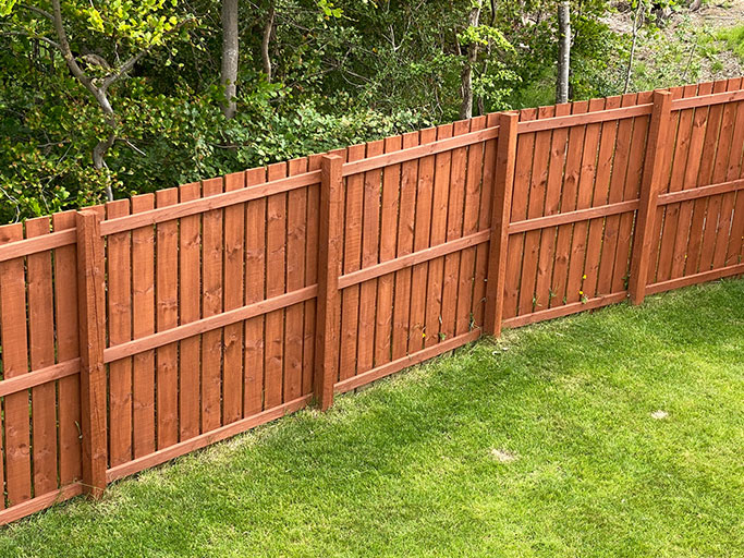 Red wooden privacy fence along a green backyard lawn with trees and shrubs beyond